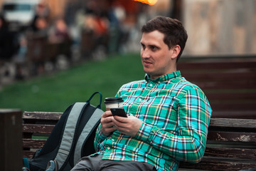 portrait of young handsome man sitting on bench drinking coffee browsing internet via phone