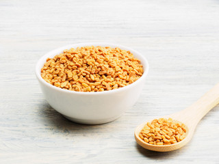 Fenugreek in white bowl and spoon on white wooden background. Indian cuisine, ayurveda, naturopathy, modern apothecary concept