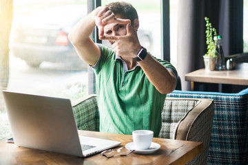 Young serious businessman in green t-shirt sitting with laptop and looking at camera with focus crop composition gesture. business and freelancing concept. indoor shot near big window at daytime.