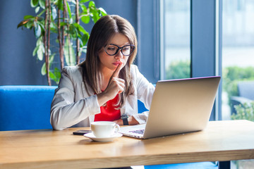 Be quiet. Portrait of serious beautiful stylish brunette young woman in glasses sitting, looking at her laptop display with finger on lips on video call. indoor studio shot, cafe, office background.