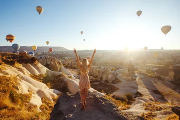 Poster Jong aantrekkelijk meisje in een hoed staat op de berg met vliegende luchtballonnen op de achtergrond. Vinger wijzend meisje in de zonsopgang. Uitzicht vanaf de achterkant. Beroemde toerist Turkse regio cappadocië. Gorem. © maxbelchenko