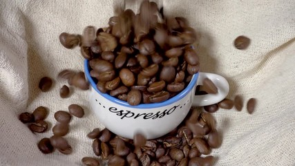 Slow motion close POV overhead shot of fresh whole coffee beans pouring in and around an overflowing espresso cup, in a hessian sack. - Powered by Adobe