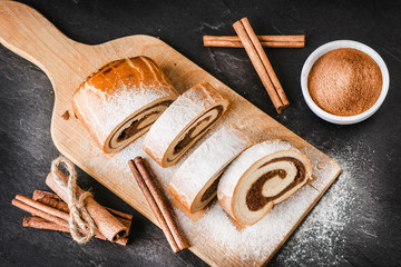 Fresh baked cinnamon strudel on dark stone table sugar covered. Tasty roll cake on cut board with black background top view.
