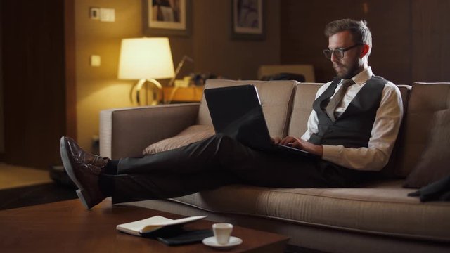 Young businessman in glasses working at a laptop, man in the suit sitting on the couch in the hotel, business trip.