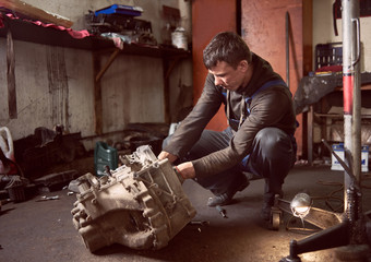 Auto mechanic squatting working on car detail on floor at auto repair shop. Strong repairman in dirty workwear in action. Tools, spare parts and garage interior on background