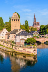 Close-up on the Heinrichsturm tower and the Ponts Couverts (covered bridges) on the river Ill in the Petite France historic quarter in Strasbourg, France, with Notre-Dame cathedral in the distance. - obrazy, fototapety, plakaty
