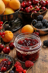 Jar of cherry jam among fresh fruits on wooden table.