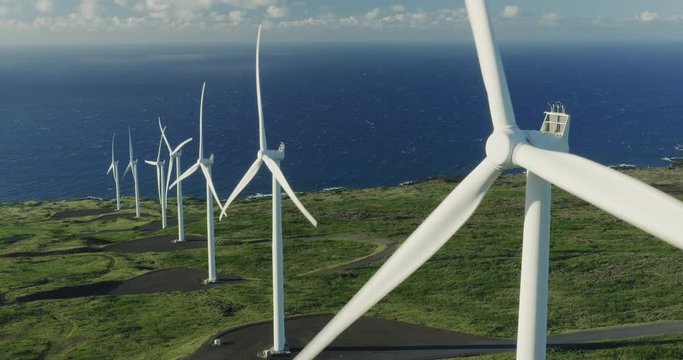 Aerial view of a row of windmills spinning surrounded by green fields and ocean in the background, view of a wind farm generating electricity, clean green energy future concept