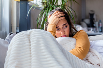 Sick woman with headache sitting under the blanket. Sick woman with seasonal infections, flu, allergy lying in bed. Sick woman covered with a blanket lying in bed with high fever and a flu, resting. - obrazy, fototapety, plakaty