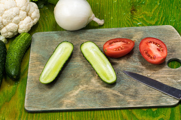 fresh vegetables, halves of tomato, cucumber, onion on a cutting board with a knife on a green wooden surface