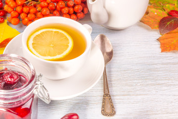 Autumn tea party - a cup of tea with lemon and a teapot on a table with autumn leaves and rowan berries and cornel jam, copy space