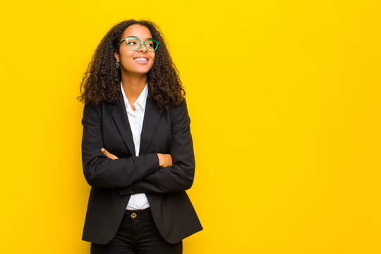 Black Business Woman Feeling Happy, Proud And Hopeful, Wondering Or Thinking, Looking Up To Copy Space With Crossed Arms Against Orange Wall