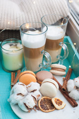 Turquoise breakfast tray with two cups of latte macchiato, macarons, cotton flowers, cinnamon rolls, dried orange slices, burning candle and string lights. Knitted sweaters on the background.