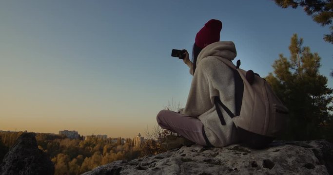 Young female sitting on a cliff at sunset doing a selfie.