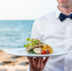 waiter holding grilled fish with lemon, tomato, creamy herbs at the seaside restaurant