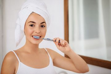 Beautiful young woman brushing teeth and looking at the camera in the bathroom. Dental health care