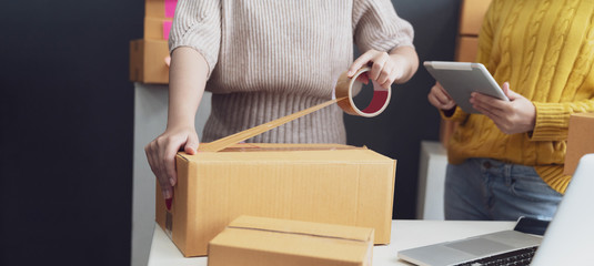 Women packing parcels with sticky tape, Entrepreneur owner using smartphone or laptop taking...
