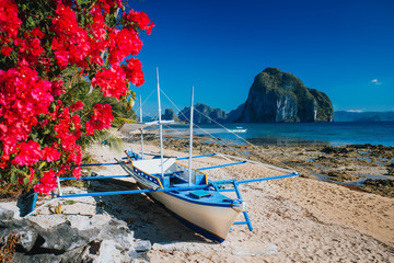 Fototapeta na wymiar Traditional banca boat and vivid colored flowers at Las cabanas beach with amazing Pinagbuyutan island in background. Exotic nature scenery in El Nido, Palawan, Philippines