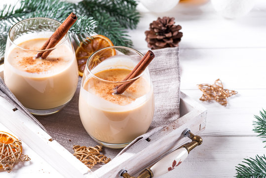 Eggnog in glasses with cinamon in tray, dry orange,fir branches and pine cone on white wooden table