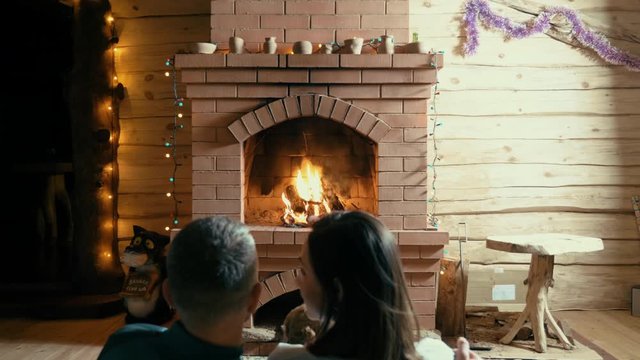 Couple and their dog sitting by brightly burning flame in fireplace decorated with festive garlands, stock video. Red brick fireplace in wooden house, place for family rest and romantic date