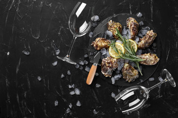 Fresh closed oysters, ice, lemon on a round slate and two empty glasses are on a black stone textured background. Top view with copy space. Close-up.