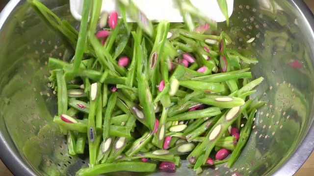 Close POV slow motion overhead shot of raw, washed and chopped, green runner beans being poured from a chopping board into a steel colander.