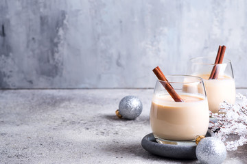 Winter spicy hot drink eggnog in a glasses with cinamon in stone plate, ice frost branches on light...