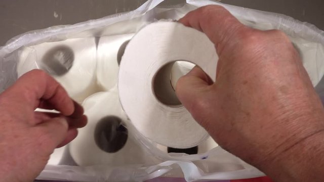 Normal speed, then slow motion: Close POV overhead shot of a man&rsquo;s hands removing a fresh white toilet roll from a multipack.