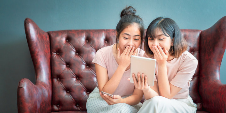 Young asian women happy couple sitting on sofa shopping online using laptop a computer and smartphone in living room at home. Two young friends watching a video on a tablet in the living room.