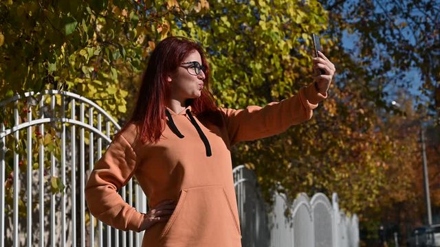 Beautiful red-haired woman in glasses makes selfie on a mobile. A happy girl in an ocher-colored hoodie is photographed on the phone by the hedge on a warm autumn day. Leaf fall.