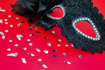 Close up of black with a feather, lace and rhinestones carnival mask with silver confetti. All on a bright red background. The concept of holiday, fun