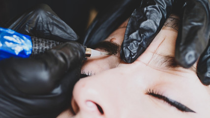 The microblading master in black gloves is filling up the shape of an eyebrow with a pigment holding with one hand woman's head, close-up, camera is above the client.