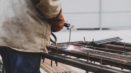 A worker is welding rebar with hollow electrode in a protective orange gloves. Sparks and smoke are allocated. Close-up side view of the process from the back of a welder. Daytime, outside.