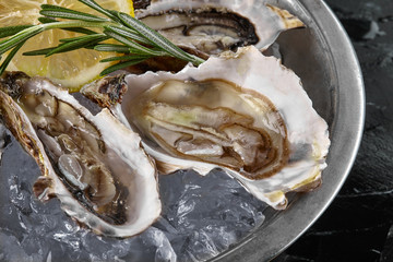 Fresh opened oysters, ice and lemon on a round metal plate, black stone textured background. Top view. Close-up shot.