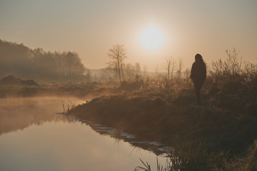 Woman walking through a meadow by a pond in the foggy morning. Sun rising above field and pond...