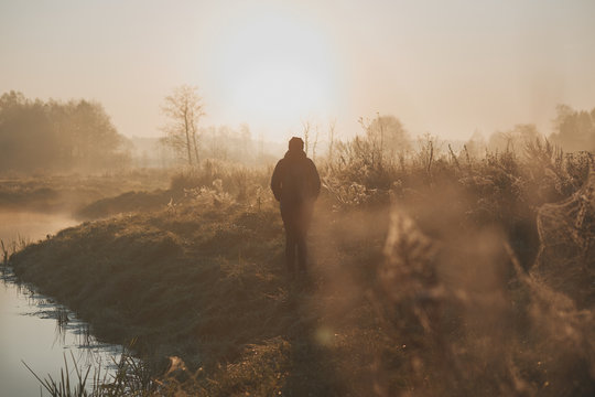 Woman walking through a meadow by a pond in the foggy morning. Sun rising above field and pond flooded with fog in the morning. Real people, authentic situations