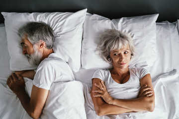 Overhead of senior couple in the bed. Pretty aged woman is angy with her sleeping husband.