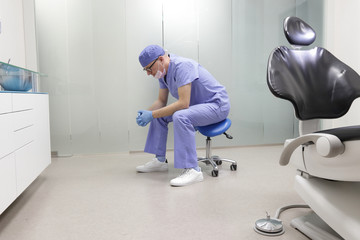 Tired middle age, male dentist in his office sitting on stool, having break,waiting for patient in...