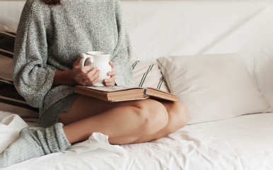 girl in home clothes on the couch reads a book and drinks a hot drink