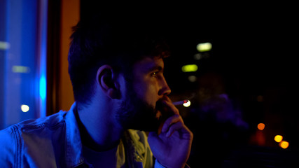 Young man smoking alone near window at night, worrying about life problems