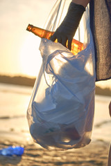 Young volunteer in black gloves is walking with garbage bag along a dirty beach of the river and cleaning up trash. People and ecology. Close-up.