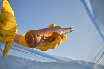 Hand of a conscious person in a yellow rubber glove puts a used brown glass bottle in a blue trash bag. Blue sky. Riverside pollution. Close-up shot.