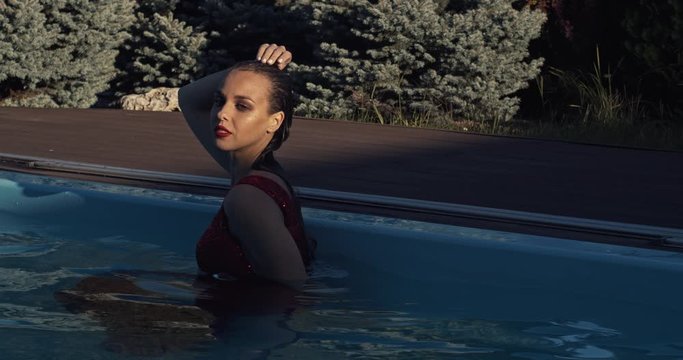 Woman with wet hair is standing in the pool in red dress, posing in 4k