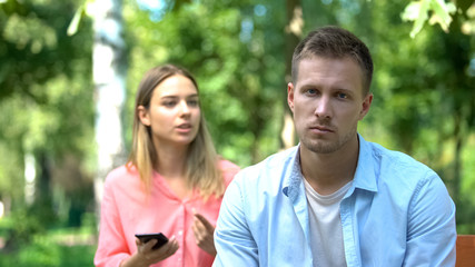 Sad young man looking camera, annoyed jealous girlfriend holding phone, distrust