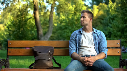 Fototapeta premium Handsome caucasian male sitting park bench with backpack, urban recreation