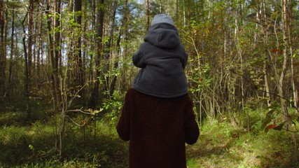 Baby sits on shoulders mother, they walk with dog in autumn forest