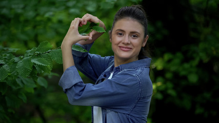 Naklejka na ściany i meble Happy woman making heart sign by hands in front of tree leaves, care of nature