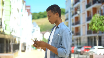 Upset mixed-race teen boy counting dollars near shop sad about purchase sum lack