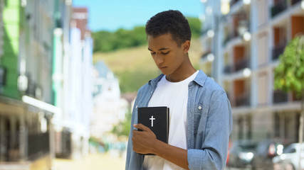 Faithful mixed-race teen male looking at holy bible in hands, religion and God