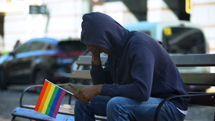 Black man in hood sitting on bench with lgbt minority flag, preconceptions